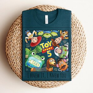 K&ouml;nnte beinhalten: Ein dunkelblaugr&uuml;nes T-Shirt mit einem Grafikdruck von Toy Story-Figuren, darunter Woody, Buzz Lightyear, Jessie, Rex, Slinky Dog, Mr. Potato Head und Aliens. Der Text lautet "TOY STORY 5", "HELLO" und "I KNEW IT, I KNEW YOU".