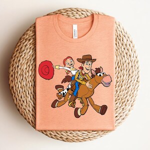 May include: Peach-colored t-shirt featuring a cartoon illustration of Woody, Jessie, and a horse from Toy Story. Woody is riding the horse, while Jessie holds a red hat. The shirt is folded on a woven surface.