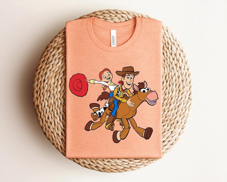 May include: Peach-colored t-shirt featuring a cartoon illustration of Woody, Jessie, and a horse from Toy Story. Woody is riding the horse, while Jessie holds a red hat. The shirt is folded on a woven surface.