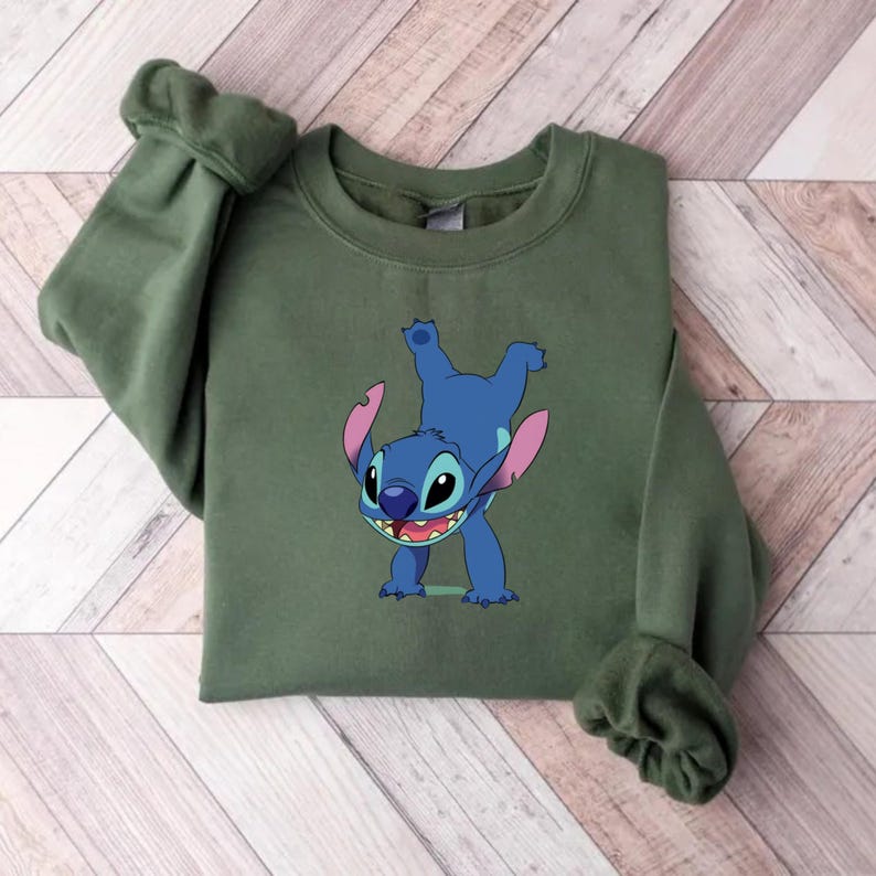 Puede incluir: Una sudadera verde oliva con un personaje de dibujos animados azul. El personaje tiene orejas grandes y una amplia sonrisa, en una pose juguetona con los brazos y las piernas en alto. La sudadera tiene mangas largas y cuello redondo.