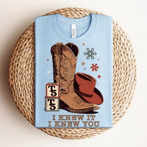 May include: A light blue t-shirt featuring a graphic of a brown cowboy boot, a red cowboy hat, two stacked blocks with the letter T and the number 5, and small flower and star icons. The text I KNEW IT I KNEW YOU is printed at the bottom.