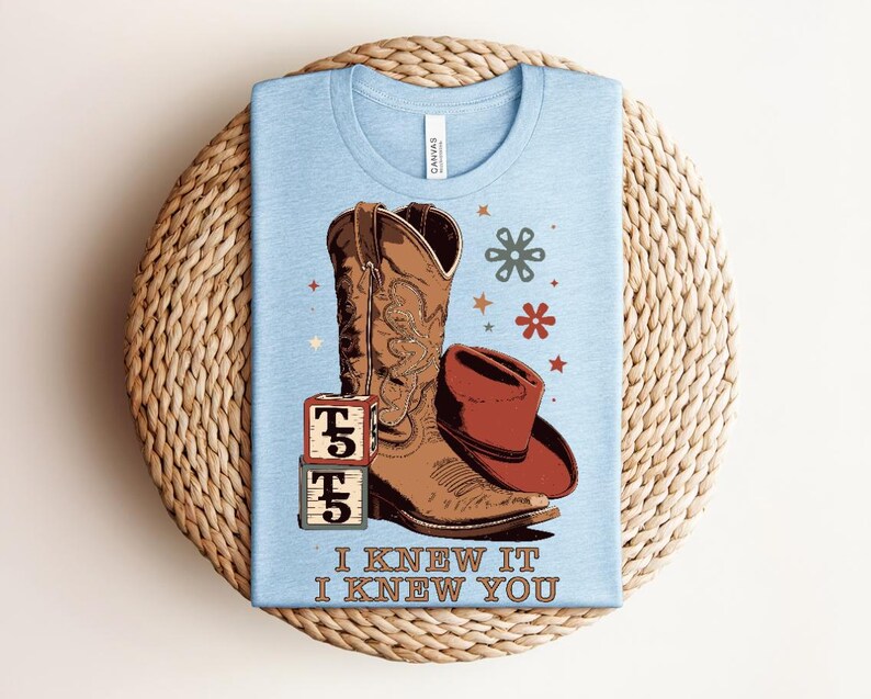 May include: A light blue t-shirt featuring a graphic of a brown cowboy boot, a red cowboy hat, two stacked blocks with the letter T and the number 5, and small flower and star icons. The text I KNEW IT I KNEW YOU is printed at the bottom.
