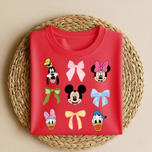 May include: A red crew neck sweatshirt with cartoon character faces and bows. Characters include Mickey Mouse, Minnie Mouse, Donald Duck, Daisy Duck, and Goofy. The bows are in various colours, including pink, green, and yellow.