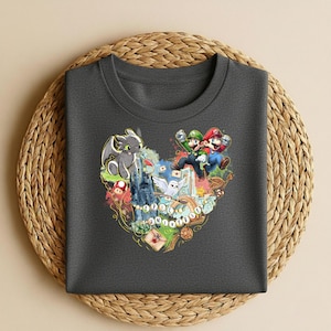 May include: Dark gray t-shirt featuring a colorful heart-shaped graphic with characters from How to Train Your Dragon, Super Mario, and Harry Potter. The graphic includes the text "EPIC ADVENTURES". The shirt is displayed on a woven, circular mat.