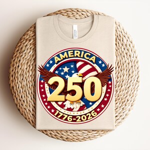 May include: A beige t-shirt with a circular design. The design features the word "AMERICA" in blue, red, and white, a golden "250", and an eagle with outstretched wings. The dates "1776-2026" are also displayed on the shirt.