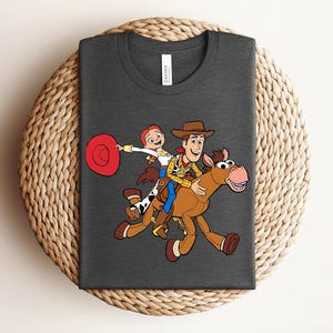 May include: Dark gray t-shirt featuring a colorful cartoon graphic of Woody, Jessie, and a horse. The characters are riding the horse, with Jessie holding a red hat. The design is centered on the shirt.