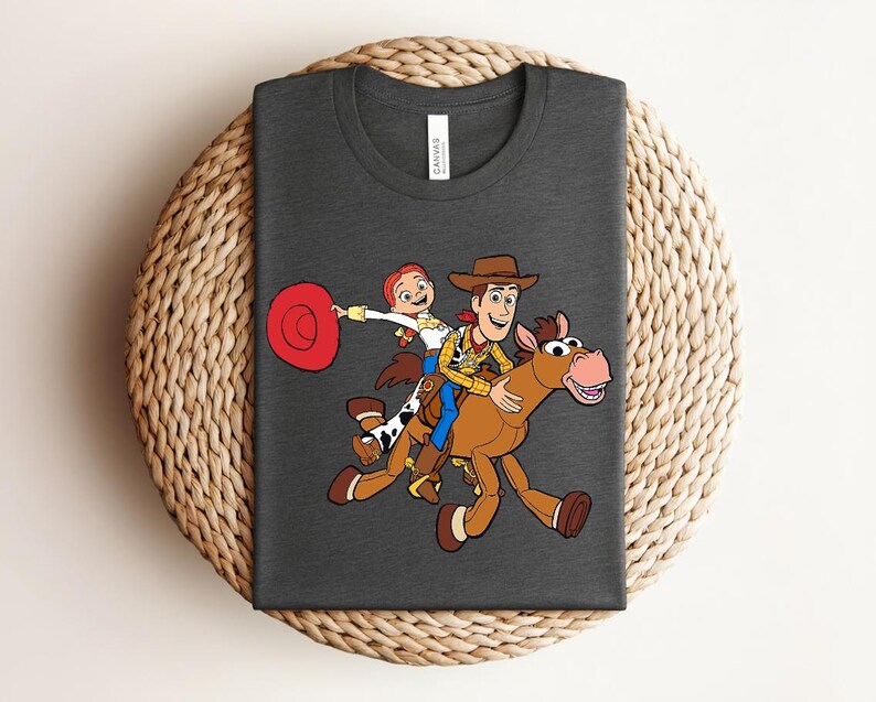May include: Dark gray t-shirt featuring a colorful cartoon graphic of Woody, Jessie, and a horse. The characters are riding the horse, with Jessie holding a red hat. The design is centered on the shirt.