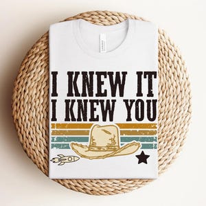 May include: A white t-shirt featuring the black text I KNEW IT I KNEW YOU above a graphic of a tan cowboy hat, a small rocket, and a black star, all set against horizontal stripes in muted shades of yellow, orange, teal, and dark green.
