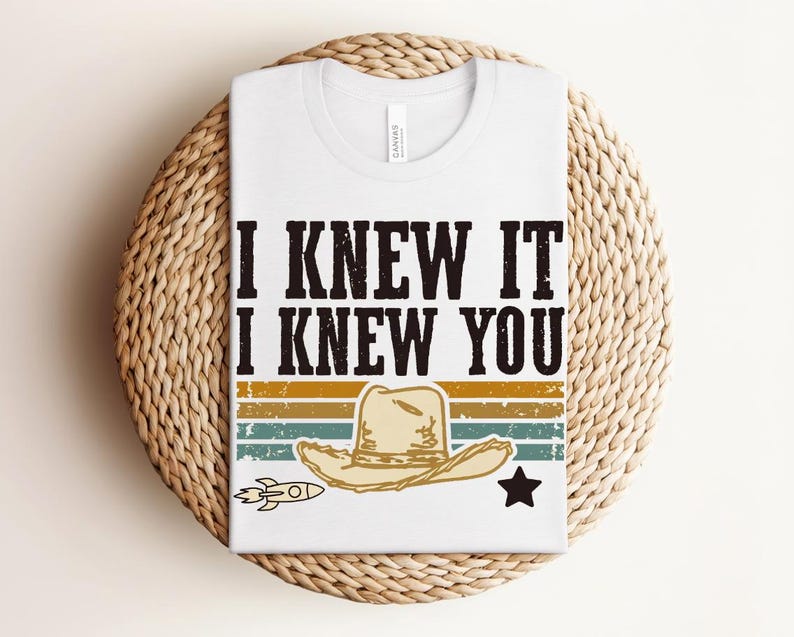 May include: A white t-shirt featuring the black text I KNEW IT I KNEW YOU above a graphic of a tan cowboy hat, a small rocket, and a black star, all set against horizontal stripes in muted shades of yellow, orange, teal, and dark green.