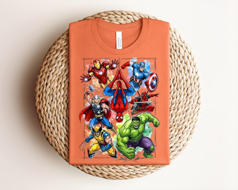 May include: Orange t-shirt with a vibrant graphic of Marvel superheroes such as Iron Man, Captain America, and Spider-Man. The shirt has a crew neck and a "Canvas" label. The design is set within a rectangular frame.