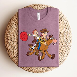 May include: Mauve t-shirt featuring a cartoon illustration of Woody and Jessie from Toy Story riding a horse. Woody wears a cowboy hat and vest. Jessie holds a red hat. The horse is brown with a pink mouth.
