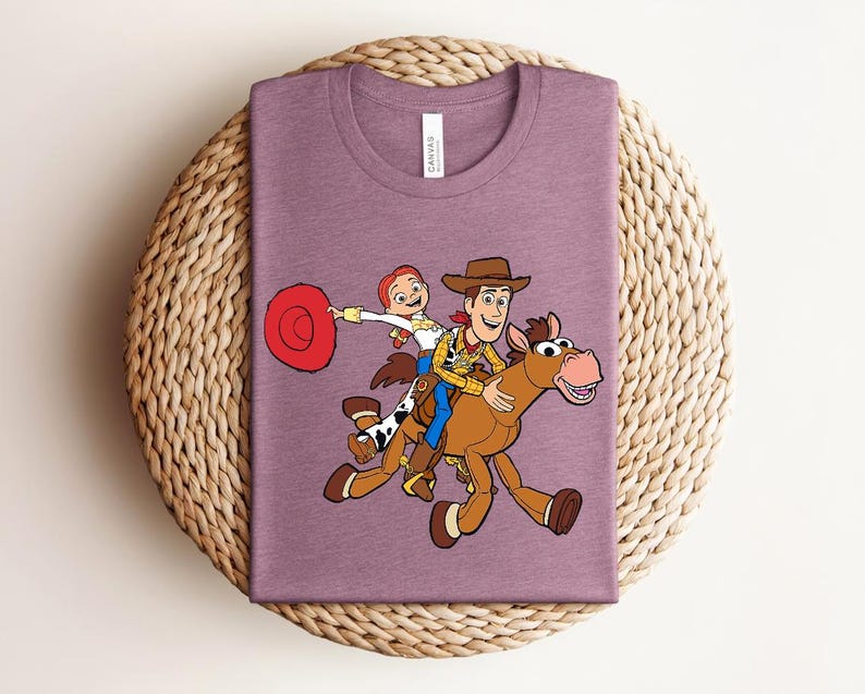 May include: Mauve t-shirt featuring a cartoon illustration of Woody and Jessie from Toy Story riding a horse. Woody wears a cowboy hat and vest. Jessie holds a red hat. The horse is brown with a pink mouth.