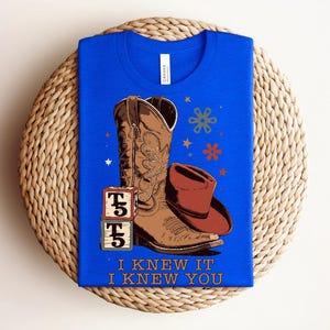 May include: A bright blue t-shirt featuring a graphic of a brown cowboy boot, a brown cowboy hat, and two stacked alphabet blocks with the letter T and number 5. The design includes small stars and flowers, with the text I KNEW IT I KNEW YOU.