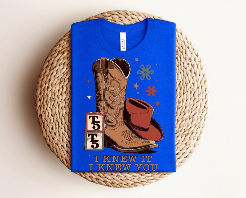 May include: A bright blue t-shirt featuring a graphic of a brown cowboy boot, a brown cowboy hat, and two stacked alphabet blocks with the letter T and number 5. The design includes small stars and flowers, with the text I KNEW IT I KNEW YOU.