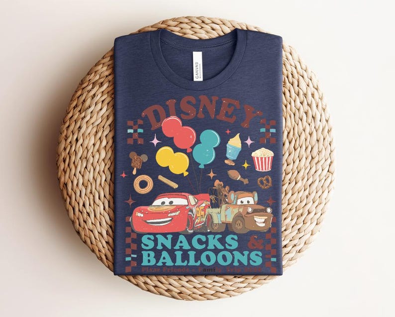 May include: Navy blue t-shirt featuring a colorful graphic with the text "Disney Snacks & Balloons." The design includes cartoon cars, balloons, and various snack illustrations. The shirt is displayed on a woven, circular surface.
