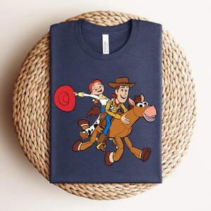 May include: Navy blue t-shirt featuring a colorful cartoon illustration of Woody, Jessie, and a horse from Toy Story. The design shows the characters riding the horse. The shirt is displayed on a woven, circular surface.