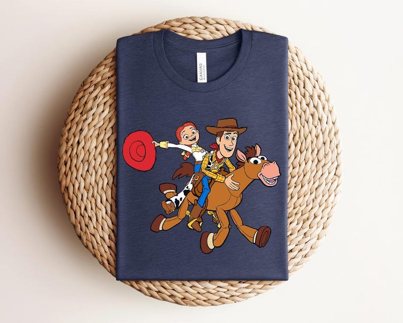 May include: Navy blue t-shirt featuring a colorful cartoon illustration of Woody, Jessie, and a horse from Toy Story. The design shows the characters riding the horse. The shirt is displayed on a woven, circular surface.