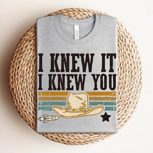 May include: A heather gray crew neck t-shirt featuring the black text I KNEW IT I KNEW YOU above a graphic of a beige cowboy hat, a small rocket, and a black star, set against horizontal stripes in muted yellow, orange, and teal colors.