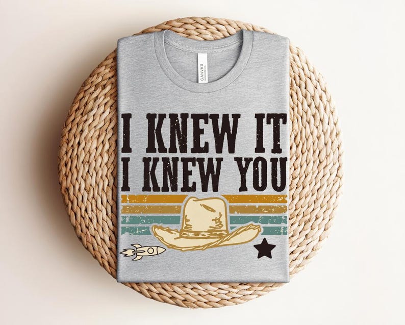 May include: A heather gray crew neck t-shirt featuring the black text I KNEW IT I KNEW YOU above a graphic of a beige cowboy hat, a small rocket, and a black star, set against horizontal stripes in muted yellow, orange, and teal colors.