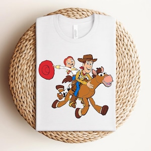 May include: White t-shirt featuring a colorful graphic of Woody and Bullseye from Toy Story. Woody is riding Bullseye, with Jessie holding a red lasso. The design is centered on the shirt.