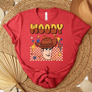 Puede incluir: Camiseta roja con un dise&ntilde;o de personaje de vaquero de dibujos animados y el nombre "WOODY" en marr&oacute;n y amarillo. El dise&ntilde;o incluye un sombrero de vaquero, estrellas y un patr&oacute;n de cuadros.