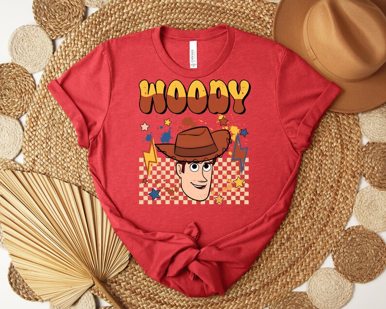 Puede incluir: Camiseta roja con un dise&ntilde;o de personaje de vaquero de dibujos animados y el nombre "WOODY" en marr&oacute;n y amarillo. El dise&ntilde;o incluye un sombrero de vaquero, estrellas y un patr&oacute;n de cuadros.