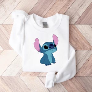 K&ouml;nnte beinhalten: Wei&szlig;es Sweatshirt mit einer Illustration einer blauen Cartoonfigur. Die Figur hat gro&szlig;e Ohren und einen freundlichen Ausdruck. Die blaue Figur hat gro&szlig;e schwarze Augen und rosa Ohren. Das Sweatshirt ist aus weichem Material und f&uuml;r den Freizeitgebrauch geeignet.