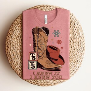May include: A dusty rose t-shirt featuring a graphic of a brown cowboy boot, a brown cowboy hat, and two stacked alphabet blocks with the letter T. The text I KNEW IT I KNEW YOU is printed below the graphic, along with small star and flower icons.