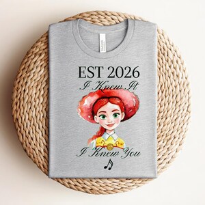 Pode incluir: Uma t-shirt cinzenta clara com uma ilustra&ccedil;&atilde;o em aguarela de uma personagem feminina de cabelo ruivo e um grande chap&eacute;u vermelho. O texto diz EST 2026, I Knew It e I Knew You, com um pequeno s&iacute;mbolo de nota musical preta abaixo.