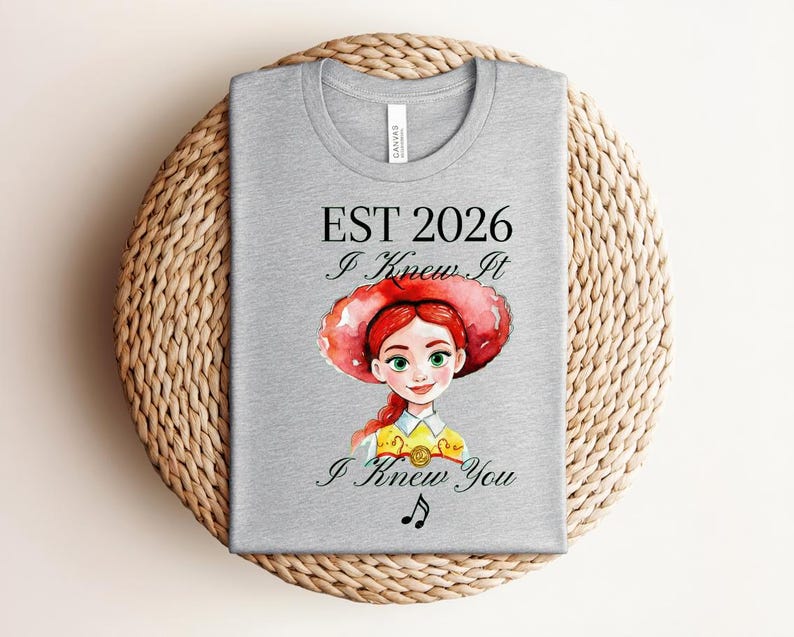 Pode incluir: Uma t-shirt cinzenta clara com uma ilustra&ccedil;&atilde;o em aguarela de uma personagem feminina de cabelo ruivo e um grande chap&eacute;u vermelho. O texto diz EST 2026, I Knew It e I Knew You, com um pequeno s&iacute;mbolo de nota musical preta abaixo.