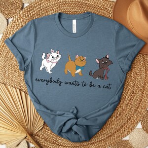 May include: A blue t-shirt featuring three cartoon cats with the text "everybody wants to be a cat". The cats are white, tan, and black, each with unique accessories. The shirt is displayed on a woven surface.