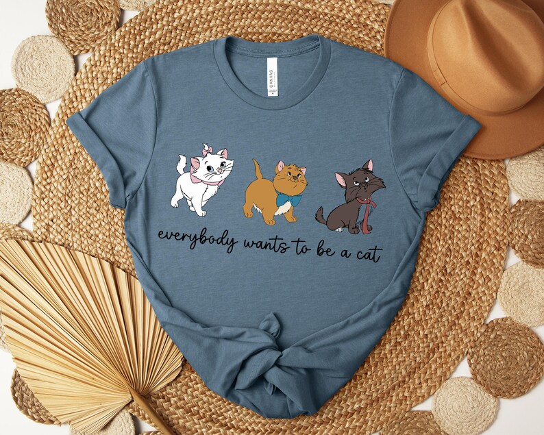 May include: A blue t-shirt featuring three cartoon cats with the text "everybody wants to be a cat". The cats are white, tan, and black, each with unique accessories. The shirt is displayed on a woven surface.