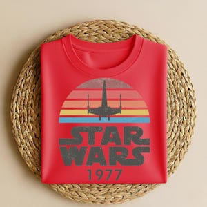 May include: A red t-shirt featuring a retro design with a silhouette of a starfighter against a sunset of orange, yellow, and blue stripes. The words "STAR WARS" are printed in a distressed font, with "1977" below.