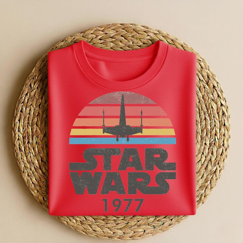 May include: A red t-shirt featuring a retro design with a silhouette of a starfighter against a sunset of orange, yellow, and blue stripes. The words "STAR WARS" are printed in a distressed font, with "1977" below.