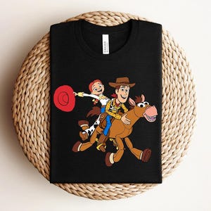 May include: Black t-shirt featuring a colorful graphic of Woody, Jessie, and a horse from the Toy Story movie. The design includes a red hat and the characters are riding the horse. The shirt is displayed on a woven surface.