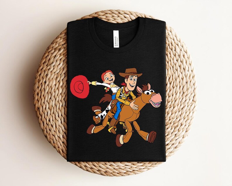 May include: Black t-shirt featuring a colorful graphic of Woody, Jessie, and a horse from the Toy Story movie. The design includes a red hat and the characters are riding the horse. The shirt is displayed on a woven surface.