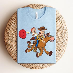 May include: Light blue t-shirt featuring a cartoon illustration of Woody and Jessie from Toy Story riding a horse. The design includes a red hat, brown cowboy hat, and a brown horse with a pink nose.