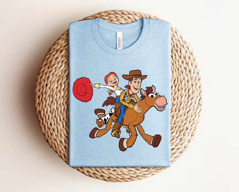 May include: Light blue t-shirt featuring a cartoon illustration of Woody and Jessie from Toy Story riding a horse. The design includes a red hat, brown cowboy hat, and a brown horse with a pink nose.