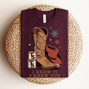 May include: A maroon t-shirt featuring a graphic of a brown cowboy boot, a red cowboy hat, two stacked alphabet blocks with the text T5, and small star and flower shapes. The text I KNEW IT I KNEW YOU is printed in a tan color at the bottom.
