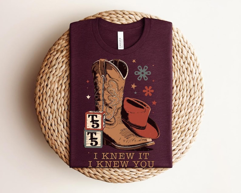 May include: A maroon t-shirt featuring a graphic of a brown cowboy boot, a red cowboy hat, two stacked alphabet blocks with the text T5, and small star and flower shapes. The text I KNEW IT I KNEW YOU is printed in a tan color at the bottom.