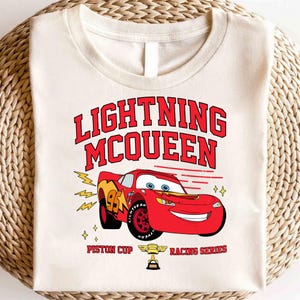 May include: A white t-shirt with a red and black graphic of Lightning McQueen from the Cars movie. The graphic includes the text "Lightning McQueen", "Piston Cup", and "Racing Series".
