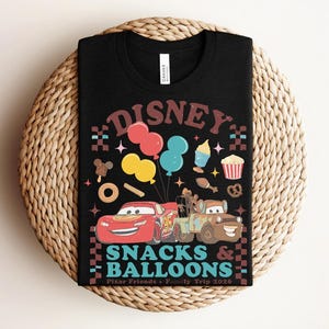 May include: Black t-shirt with a colorful graphic featuring the word "DISNEY" in brown, balloons, cartoon cars, and snacks. The text "SNACKS & BALLOONS" is below the cars, with "Pixar Friends + Family Trip 2026" at the bottom.