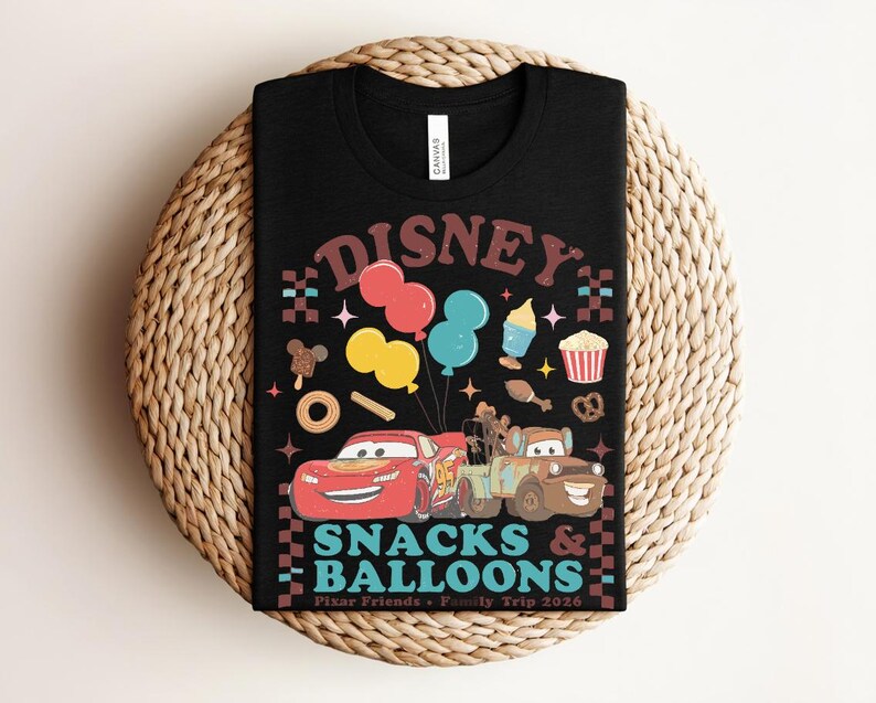 May include: Black t-shirt with a colorful graphic featuring the word "DISNEY" in brown, balloons, cartoon cars, and snacks. The text "SNACKS & BALLOONS" is below the cars, with "Pixar Friends + Family Trip 2026" at the bottom.