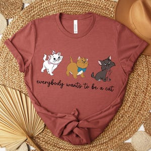 May include: A maroon t-shirt featuring three cartoon cats with the text "everybody wants to be a cat". The cats are white, tan, and gray, each with unique accessories. The shirt is folded on a woven surface.