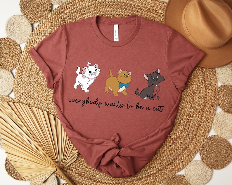 May include: A maroon t-shirt featuring three cartoon cats with the text "everybody wants to be a cat". The cats are white, tan, and gray, each with unique accessories. The shirt is folded on a woven surface.