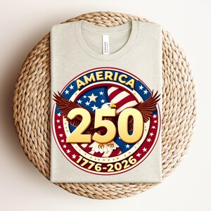 May include: A folded light grey t-shirt with a circular graphic. The graphic includes the word "AMERICA" in blue and red, the number "250" in gold, and an eagle with outstretched wings. The dates "1776-2026" are also present.