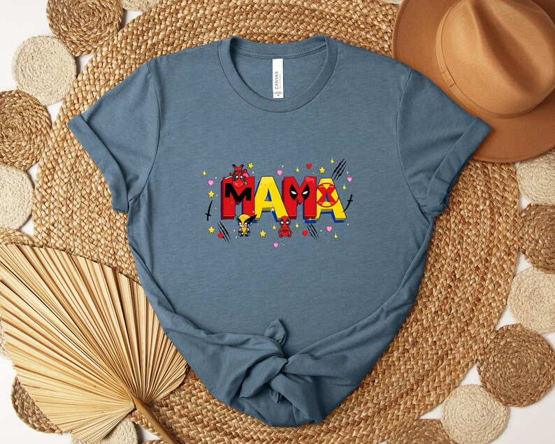 Puede incluir: Una camiseta azul con la palabra "MAMA" en rojo y amarillo, con gr&aacute;ficos tem&aacute;ticos de superh&eacute;roes. La camiseta tiene cuello redondo y mangas cortas. El dise&ntilde;o incluye corazones, estrellas e ilustraciones de personajes.