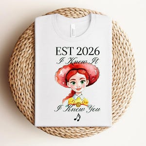 Pode incluir: Uma t-shirt branca com uma ilustra&ccedil;&atilde;o em estilo aguarela de uma personagem ruiva com um chap&eacute;u de cowgirl vermelho e uma camisa amarela. O texto diz EST 2026, I Knew It, I Knew You, acompanhado por uma pequena nota musical preta.
