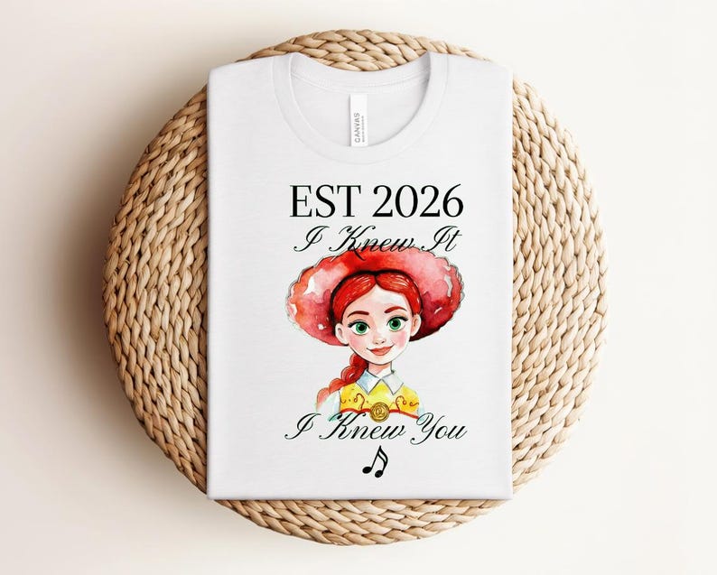 Pode incluir: Uma t-shirt branca com uma ilustra&ccedil;&atilde;o em estilo aguarela de uma personagem ruiva com um chap&eacute;u de cowgirl vermelho e uma camisa amarela. O texto diz EST 2026, I Knew It, I Knew You, acompanhado por uma pequena nota musical preta.