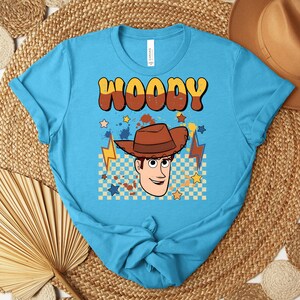 Puede incluir: Una camiseta turquesa con una ilustraci&oacute;n de dibujos animados de Woody de Toy Story. El dise&ntilde;o incluye el nombre "WOODY" en letras marrones y amarillas, una imagen de Woody, estrellas y rayos.